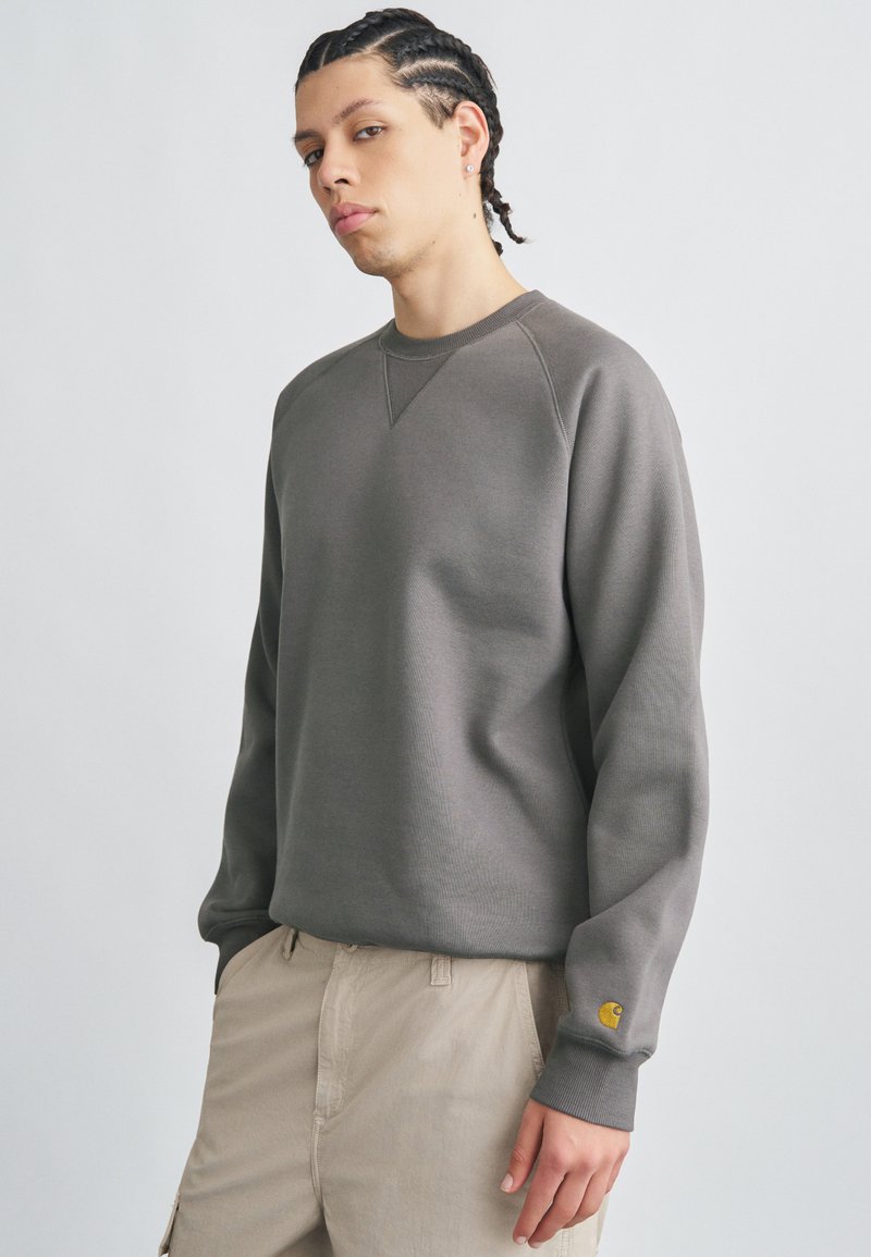Gray sweatshirt with a ribbed texture, crew neckline, and raglan sleeves. Features a small yellow logo on the cuff and a relaxed fit.