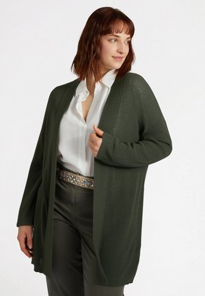 Cardigan long à ouverture frontale en vert foncé, texture côtelée. Présente une coupe ample et des manches larges, accompagnée d'un blouse blanche et d'une ceinture embellie.