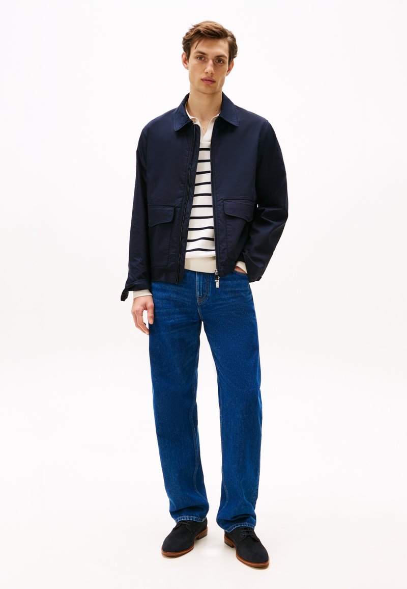 Man in een marineblauwe jas, wit gestreept shirt, blauwe spijkerbroek en zwarte schoenen, staande met één hand in zijn zak tegen een witte achtergrond.