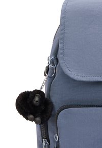 Grå tygryggsäck med klaffstängning, dragkedjefickor och en svart fluffig gorillanyckelring som är fäst på sidan.