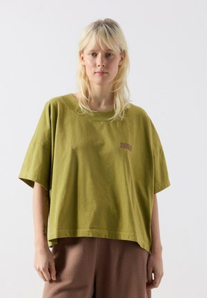 Blonde person wearing an oversized olive green T-shirt with small "American Vintage" text, standing against a plain light background.