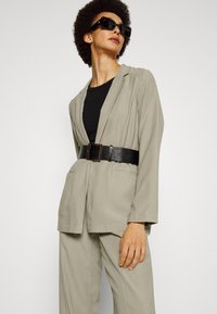 Blazer cinza claro ajustado com um cinto preto apertado. Apresenta dois bolsos frontais e é complementado por uma blusa preta por baixo.