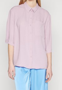 Woman wearing a loose pale pink button-up shirt with a chest pocket and short sleeves, paired with shiny light blue pants.