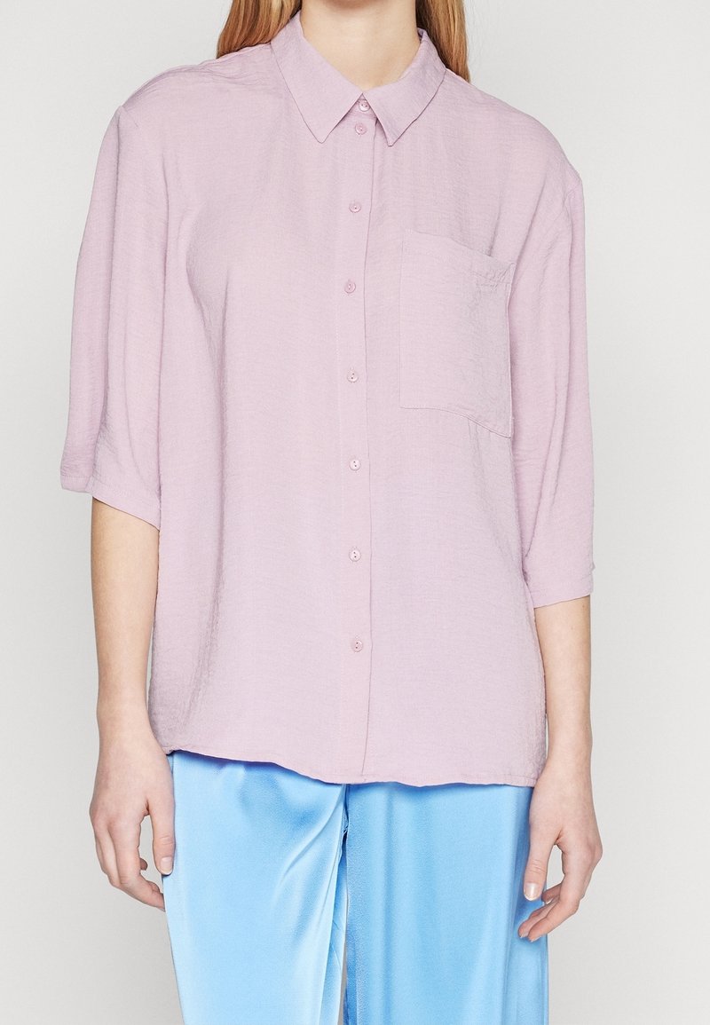 Woman wearing a loose pale pink button-up shirt with a chest pocket and short sleeves, paired with shiny light blue pants.