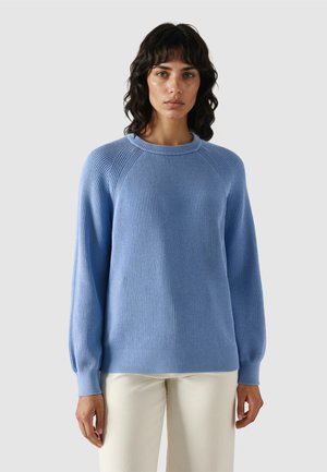 Kobieta o falujących ciemnych włosach, ubrana w jasnoniebieski prążkowany sweter i kremowe spodnie, stojąca na szarym tle.