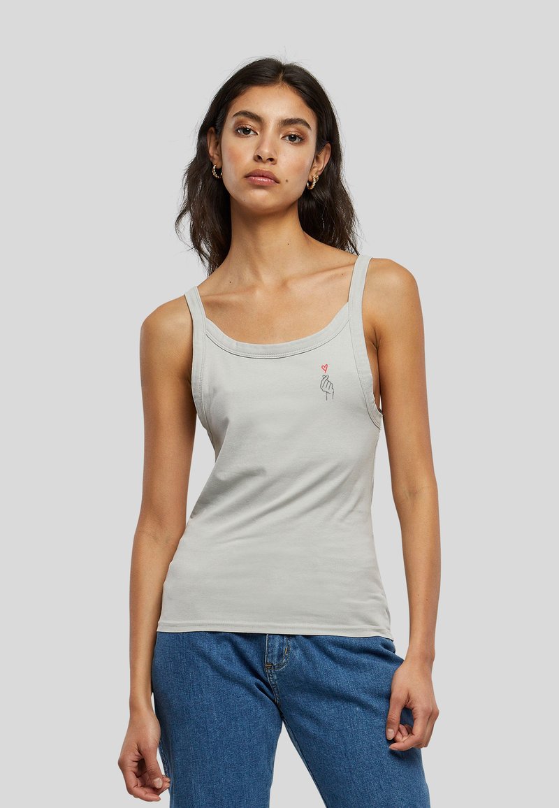 Femme portant un débardeur gris clair avec un petit motif main et cœur sur la poitrine, associé à un jean bleu, debout devant un fond uni.