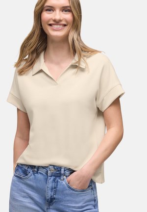 Uśmiechnięta blondynka w beżowej koszuli z krótkim rękawem i niebieskich dżinsach, z jedną ręką swobodnie w kieszeni na jednolitym tle.