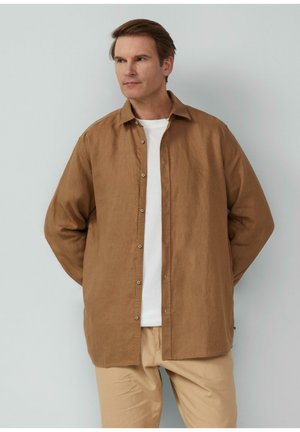 Bruine linnen overhemden met een ontspannen pasvorm, lange mouwen en een klassieke kraag. G gedragen over een wit t-shirt, gecombineerd met lichtbeige broek.