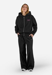 Felpa nera con zip e logo ricamato rosa, dotata di tasche a marsupio, abbinata a pantaloni larghi neri. Entrambi sono realizzati in tessuto morbido.