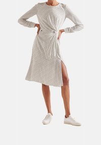 Biała sukienka z dopasowaną talią, do kolan, długimi rękawami i bocznym rozcięciem; posiada czarny, geometryczny wzór w kropki. Noszona z białymi sneakersami.