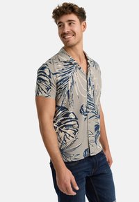 Camisa de botones de manga corta en beige, con patrones de hojas tropicales azules. Tejido ligero con un corte relajado y una textura suave.