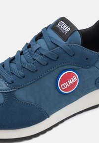 Blå sportssko med en texturerad mocka- och tygöverdel, rund röd logotypaccent och kontrasterande vit sula med svart mönster.