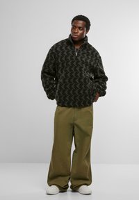 Pullover in pile nero e grigio a motivo con colletto alto e zip frontale, abbinato a pantaloni larghi verde oliva e sneakers bianche.