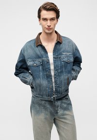 Τζιν μπουφάν σε μεσαίο μπλε με καφέ γιακά, με μπροστινές τσέπες, μεταλλικά κουμπιά και λιγότερο έντονο ξεθώριασμα. Φορεμένο πάνω από λευκό αμάνικο μπλουζάκι.