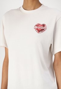 Valkoinen puuvilla t-paita, jossa on punainen sydänkuvio ja "HUGO"-teksti. Lyhyt hiha ja pyöreä kaula-aukko. Pehmeä pinta, rento istuvuus.