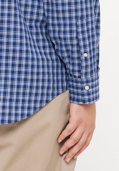 Μπλε και λευκή καρό πουκάμισο με μακριά μανίκια και κουμπωτές μανσέτες, με ομαλή υφή και στρογγυλεμένο στρίφωμα, συνδυασμένο με μπεζ παντελόνι.