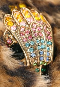 Anello a forma di mano in tonalità oro decorato con strass multicolori in rosa, blu, verde e giallo, posizionato su uno sfondo simile a pelliccia.