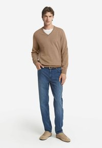 Jasnobrązowy sweter w serek, niebieskie dżinsy i beżowe buty. Ubranie ma luźny krój i gładką fakturę, bez widocznych wzorów.