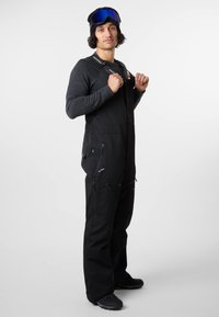 Black waterproof ski overalls with adjustable straps, multiple zippered pockets, and a relaxed fit, paired with gray long-sleeve top and goggles.