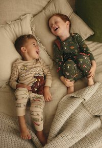 Deux enfants portent des pyjamas assortis. Le garçon à gauche a un haut à rayures beiges avec un motif graphique et un bas de pyjama beige. Le garçon à droite porte un haut vert foncé avec un motif forestier et un bas assorti. Ils sont assis sur un canapé de couleur claire avec des coussins en texture et une couverture en tricot.