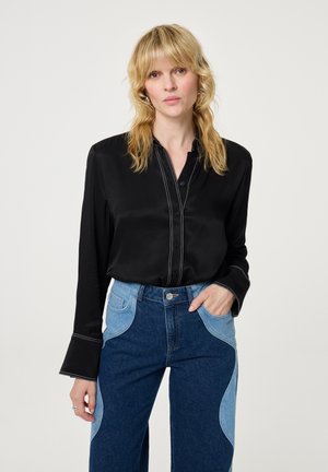 Zwart satijnen shirt met een knoopsluiting aan de voorkant en witte stiksel, gedragen met hoog-getailleerde blauwe jeans met contrasterende lichtblauwe zijpanelen.