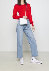 Roter Pullover mit langen Ärmeln, hellblaue gerade Jeans, weiße Umhängetasche und weiße High-Top-Sneaker, vor einem einheitlichen Hintergrund.