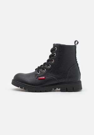 Botte de combat noire en cuir à lacets avec une semelle épaisse et une étiquette rouge Levi's sur le côté, montrée vue de l'extérieur sur fond blanc.
