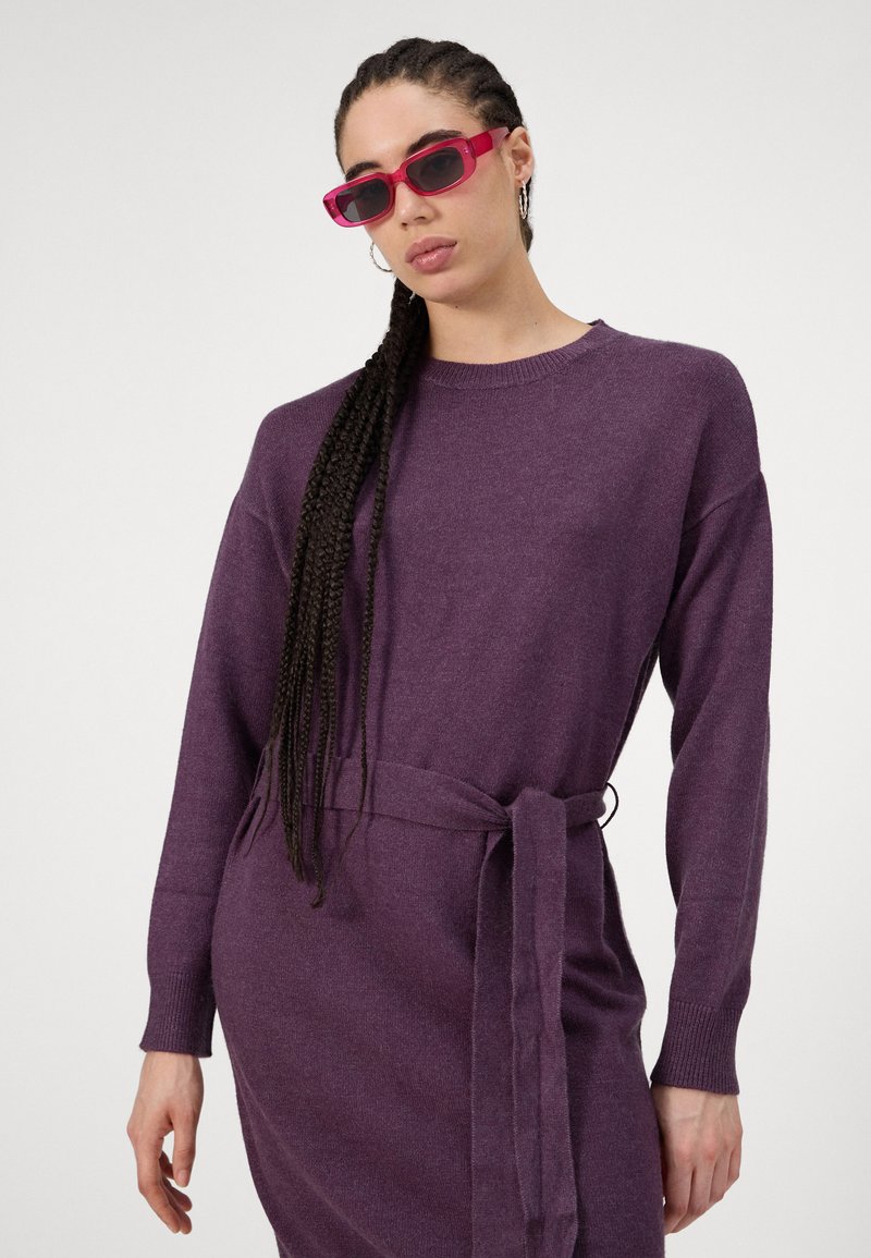 Lila Strickkleid mit langen Ärmeln und tailliertem Gürtel. Hat einen runden Ausschnitt und eine weiche Textur. Das Model trägt pinke rechteckige Sonnenbrillen.