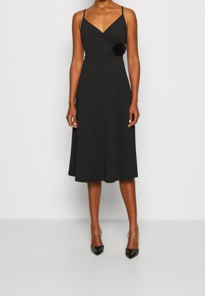 Kobieta ubrana w czarną, bezrękawnikową sukienkę midi z cienkimi ramiączkami i tkaninowym kwiatem przy talii, zestawiona z czarnymi szpiczastymi butami na obcasie.