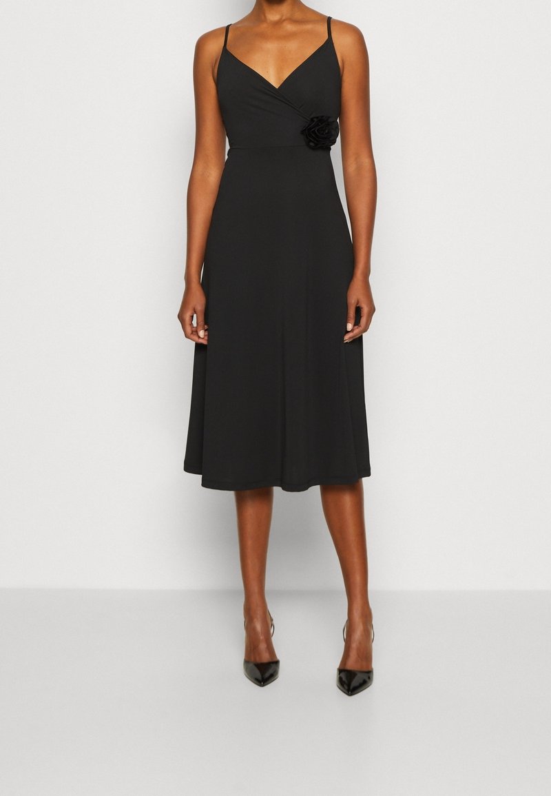 Femme portant une robe midi noire sans manches avec de fines bretelles et un détail de fleur en tissu à la taille, assortie à des talons pointus noirs.