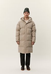 Άνδρας στέκεται φορώντας μακριά μπεζ επενδυμένη παλτό με κουκούλα, σκούρο παντελόνι, καφέ παπούτσια και σκούφο σκούρου πράσινου χρώματος με λευκό λογότυπο.