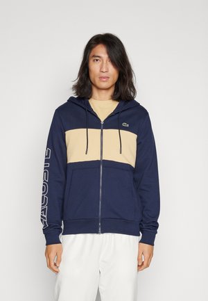 Sweat-shirt zippé bleu marine avec un panneau beige sur la poitrine, doté d'une poche kangourou, d'une capuche avec cordon de serrage et d'un texte logo blanc sur la manche.