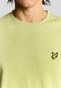 Lys grøn bomulds T-shirt med et sort og gult broderet fuglelogo på venstre brystområde; klassisk rund halsudskæring.