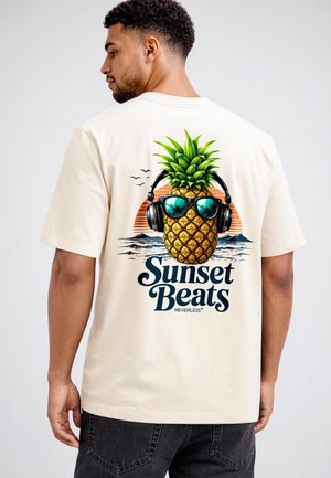 Mann trägt beige T-Shirt mit einer Grafik von einer Ananas, die Sonnenbrille und Kopfhörer trägt, mit Sonnenuntergang und Meer im Hintergrund.