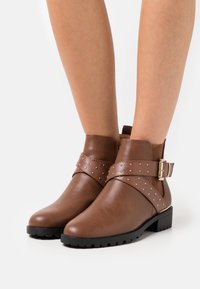 Hnědé kotníkové boty vyrobené z hladké kůže, s širokým texturovaným páskem a kovovými hroty, doplněné bočním sponou. Čtveratá špička a nízký podpatek.