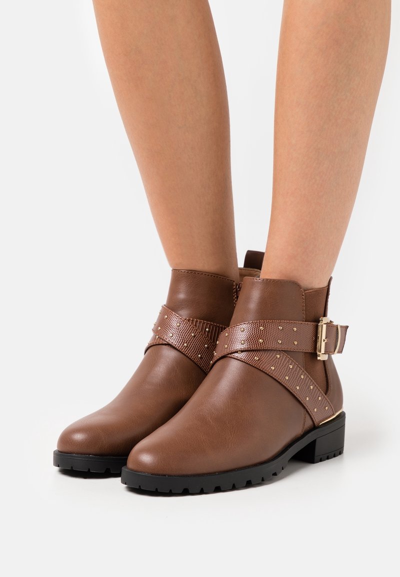 Hnědé kotníkové boty vyrobené z hladké kůže, s širokým texturovaným páskem a kovovými hroty, doplněné bočním sponou. Čtveratá špička a nízký podpatek.