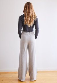 Kvinna med långt, vågigt blont hår står vänd mot en vit vägg, klädd i en svart långärmad topp och löst sittande grå byxor på ett trägolv.