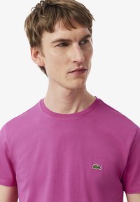 Camiseta de algodón rosa con mangas cortas, cuello redondo y logo bordado de cocodrilo verde en el lado izquierdo del pecho. Textura suave.