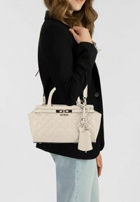 Beige doorgestikte handtas met metalen beslag en bedel, voorzien van dubbele handvatten en verstelbare riem, gedragen over de schouder bij een zwarte jas.