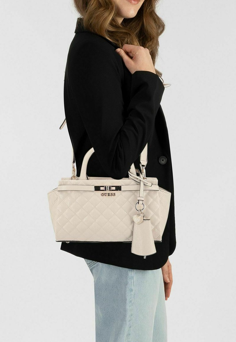 Beige doorgestikte handtas met metalen beslag en bedel, voorzien van dubbele handvatten en verstelbare riem, gedragen over de schouder bij een zwarte jas.