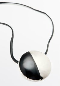 Collana con pendente rotondo caratterizzata da un design diviso in metà nero e argento, con un cordino in pelle. Il contrasto tra la superficie liscia del metallo e quella testurizzata.