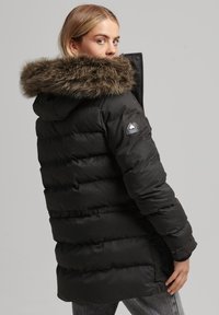 Czarna ocieplana kurtka zimowa z kapturem z sztucznego futra, z logo na rękawie oraz teksturowaną powierzchnią z poziomym przeszyciem.