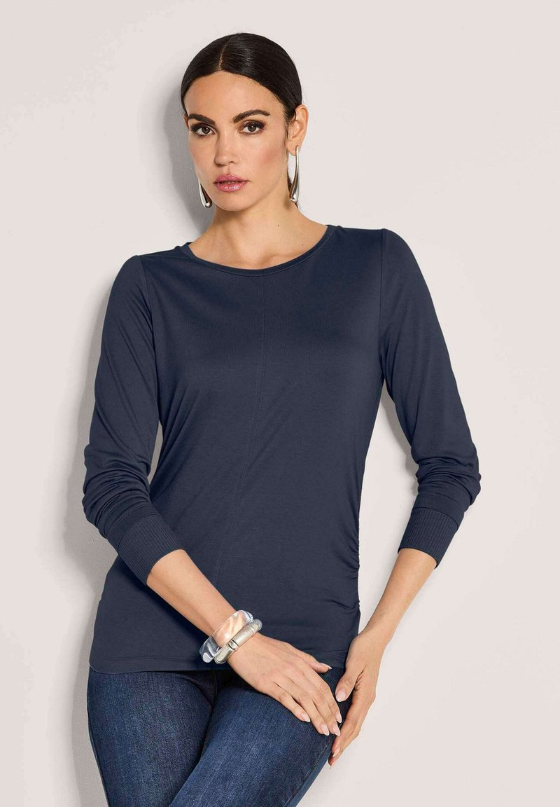 Vrouw met donker haar in een marineblauwe lange mouw top en blauwe jeans, met grote zilveren oorbellen en stevige doorzichtige en metalen armbanden.