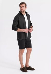 Schwarzes Hemd mit Knöpfen, darunter ein weißes T-Shirt, schwarze Shorts und braune Bootsschuhe; glattes Material, aufgerollte Ärmel, lässige Passform.