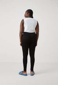 Svarta slim fit-jeans, vit ärmlös topp, flätad frisyr, bär blårandiga slip-on sandaler. Slät tygkvalitet, figurnära design, minimalistisk stil.
