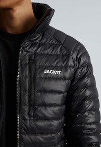 Doudoune noire à design matelassé, col montant et poche poitrine zippée. Présente le logo "JACK1T" en blanc sur le devant.