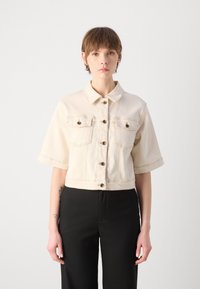 Beskarvad beige denimjacka med korta ärmar, två bröstfickor och guldtonade knappar. Har kontraststickning och en klassisk krage.