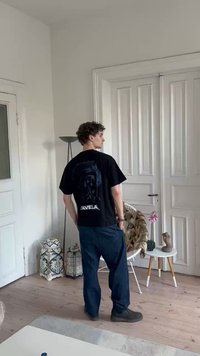 Melna grafiskā t-kreklā ar lielu piegriezumu, kas aizmugurē ir ar dizainu. Savienots ar tumši ziliem biksēm un pelēkiem apaviem, gaišā telpā.