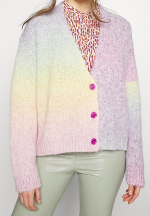 Femme portant un cardigan en maille dégradé pastel avec des boutons roses sur un chemisier à motifs et un pantalon en cuir vert clair.