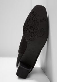 Semelle de chaussure noire avec motifs ronds de pièces en relief, texte "KAISER" et taille 41 1/2 visibles sur un fond de surface lisse.